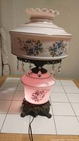 Vintage floral hurricane table lamp on tiled surface under bright light showing pink glass with floral design and decorative hanging crystal prisms.