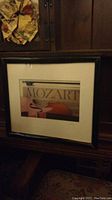 Full view of the framed Mozart wall art piece placed on a wooden surface, showing the dark frame and cream matting around the art.
