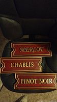 Front view of three wooden wine plaques with red background and gold text: Merlot, Chablis, Pinot Noir.