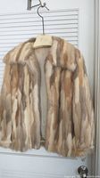 Front view of a women's genuine fur coat in shades of beige, brown, and cream hung on a silk hanger, showing overall pattern and collar.