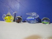 Front view of all four vintage glass paperweights in the lot, showing a teapot-shaped Joe St.Clair art glass, two spherical Millefiori style paperweights, and a bunny-shaped Millefiori style paperweight.