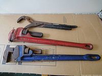 Photo of all three tools together laid flat on cardboard surface: red Reed pipe wrench, blue Kobalt pipe wrench, and black Husky groove joint plier.
