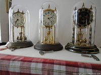 Full view of the three Kundo anniversary 400-day clocks together, showing glass domes, bases in silver, gold, and black, and three clock keys.