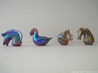 Four iridescent glass animal figurines displayed side by side against a plain background: horse head, swan, pig, and bird head, showing overall set.