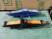 Photo showing both blue and black scissor-style car jacks side by side on green surface, without handles.