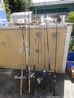 Nine fishing rods standing upright leaning against a beige metal cabinet outdoors, three have reels attached, one rod missing its tip, rods show signs of use and dirt.