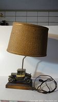 Full view of the vintage car lamp showing brown fabric lampshade, metal car model, wood base, and power cord with switch.
