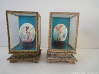 Pair of vintage hand-painted decorative eggs within glass display cases on wooden stands, showing overall presentation and condition.