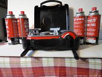 Portable Coleman single burner butane stove in an open black carrying case surrounded by four unopened Coleman butane fuel cans with red caps.