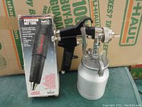 Milwaukee precision hot tool in original box next to Central spray gun with white container, all on a cardboard U-Haul box background.