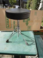 Round adjustable drum throne stool with black cushion seat and metal tripod base on plastic table against cardboard background.