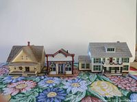 Three handcrafted miniature buildings from the Sarah Plain and Tall collection displayed on a floral fabric surface against a white background.