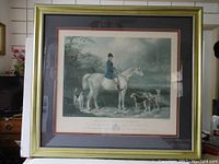Framed colored lithograph showing a rider on a white horse with dogs and outdoor background under glass within a decorative frame.