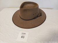 Brown felt wide-brimmed Australian cattleman style hat with braided leather band and tassels, shown on white cloth background.