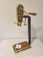 Full view of the brass and metal Le Grape bottle opener mounted on its stand, showing the wooden handle, metal arm, and base with wooden panel insert.
