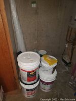 Several large buckets of paint and primer, some labeled CIL Premium and CertainTeed Mold Defense Primer, stacked in a storage area with some tools on top.