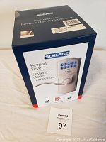 Front panel of closed blue and white box with image of Schlage keypad lever lock in satin nickel finish, showing numeric keypad and lever handle.