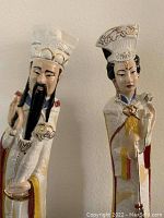 Front view of two tall Asian ceramic figurines showing detailed traditional dress, facial features, and hand-painted designs.