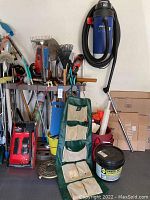 Overall view of the gardening rack with various hand tools and the green fabric hanging garden tool organizer, 5-gallon bucket of polymeric sand on floor, boxed item in background