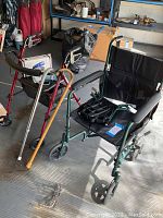 Photo showing the green folding wheelchair, red walker with padded seat, two canes, and blood pressure monitor box in the background