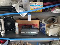 Shelf with two Oreck portable vacuums in box and cleaner spray can, plus part of a white appliance with hose