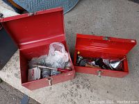 Two red metal toolboxes placed open on concrete surface showing contents inside including tools and hardware items.