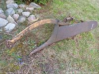 Side angle view of the antique horse drawn plow showing the long handle and curved plow blade resting on grass near rocks and shrubs.