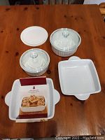 Set of four baking ware items on wooden table: two round Corning casserole dishes with glass lids, one rectangular PC baking dish with ribbed sides, and one white sealing lid.