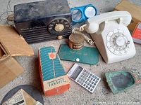 Photo showing all items spread out: rotary phone, tube radio, alarm bell, metronome, swim goggles, calculator, tin container, paper booklet, ashtray.