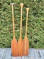Full view of three vintage cedar canoe paddles standing side by side against a hedge background on a wooden deck.