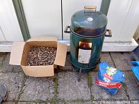 Full view of green Coleman vertical smoker with wooden handle on lid, box of apple wood chunks, and bag of charcoal placed on ground outside.