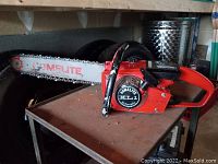 Side angle showing full length of chainsaw with blade and chain on a metal surface with tires in background.