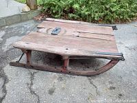 Side view of antique log sled showing curved wooden runners with metal reinforcements and wooden plank base.