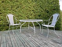 Full view of vintage Hauser patio set on wood deck, showing round glass-top table with two matching white painted metal mesh chairs.