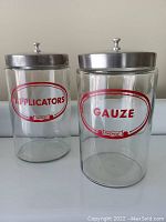 Two glass jars labeled 'APPLICATORS' and 'GAUZE' with stainless steel lids against a neutral background.