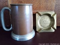 Brass ashtray and copper tankard shown side by side on brown surface with wood background.