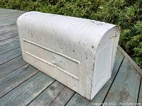 Side view of the off-white rounded galvanized mailbox showing general condition and exterior details including ribbed metal and wood base.