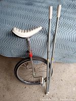 Photo of the unicycle showing wheel, frame, adjustable seat, and two metal training poles leaning against a surface