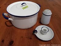 Photo showing large white enamel pot with blue trim and lid, white enamel candle holder, and muffin shaker on wooden surface.