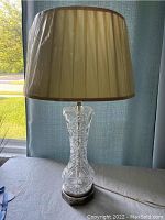 Full view of vintage cut glass lamp with beige pleated fabric shade placed on table in front of window.
