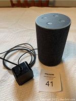 Amazon Echo smart speaker with four buttons on top and dark fabric exterior, placed upright on a light textured surface with attached black power adapter beside it.