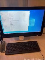 Front view of Dell all-in-one desktop powered on displaying Windows desktop with Dell logo on bezel and front stand with attached wired keyboard.