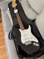 Full view of black Squier Stratocaster electric guitar with white pickguard and included soft gig bag on couch