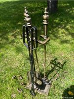 Full view of metal fireplace tools set on green grass, showing five tools with decorative finials on a sturdy stand