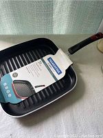 Full view of black 11 inch square grill pan with ridges and attached handle, with original label showing brand and size.