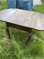 Front and side view of vintage wooden drop leaf table with both leaves extended, showing wear and surface discoloration, set outdoors on grass.