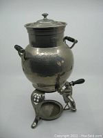 Full view of the antique WMF coffee maker showing hammered metal body, wooden side handles, spout with wooden lever, and tripod stand with drip tray.