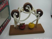 Wooden puzzle with three posts, three wooden rings threaded on ropes, and two colored wooden balls (red and yellow) attached to ropes on a wooden base.