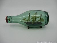 Side view of the green glass bottle containing the detailed ship model with tan sails and a red seal on the neck, showing the item in entirety.