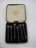 Open display case showing six sterling silver condiment spears with detailed rooster-shaped handles on black velvet lining.